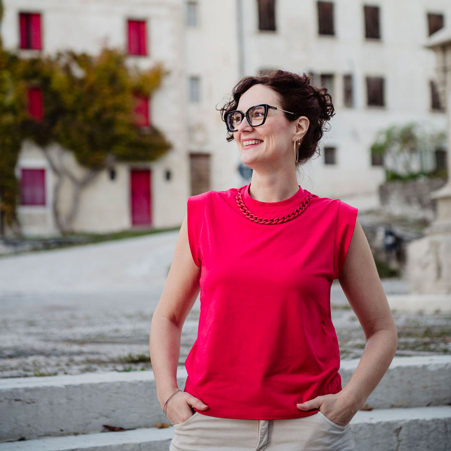 Ritratto di Irene Rizzi che indossa una maglietta color lampone, e guarda spensierata verso sinistra, sullo sfondo ci sono una piazza e dei palazzi